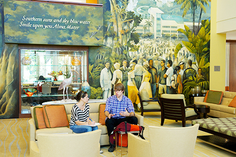 A student speaks with someone, who writes down notes on a clipboard. This is in the alumni center, an area with a painted mural in the background of people in South Florida University of Miami. There is a display case full of glass art in the back. A sculpture of an Ibis sits on a table behind the couch the two people are sitting upon. 